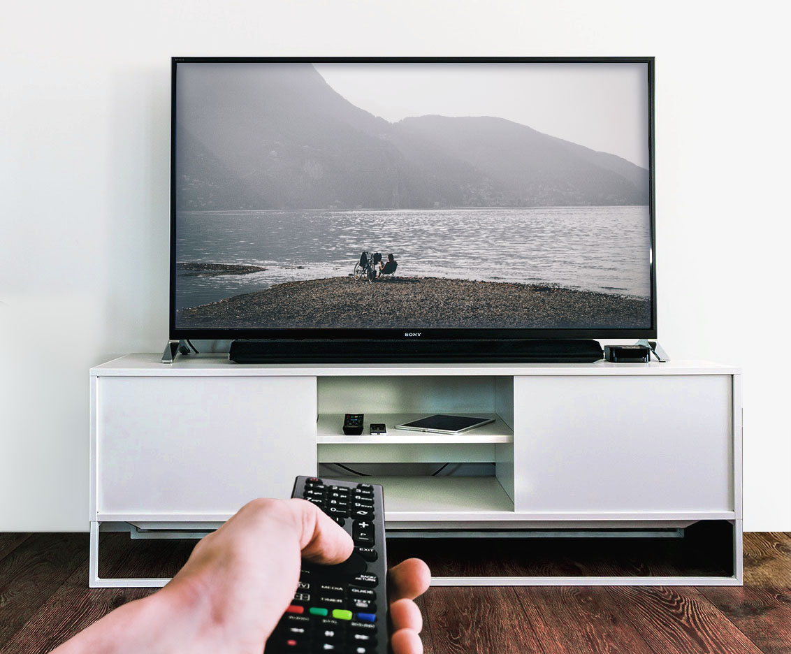 Una mano sosteniendo un control remoto negro apuntando hacia un televisor de pantalla plana que muestra una fotografía de un lago rodeado de montañas, colocado sobre un mueble de televisión blanco en una habitación con piso de madera.
