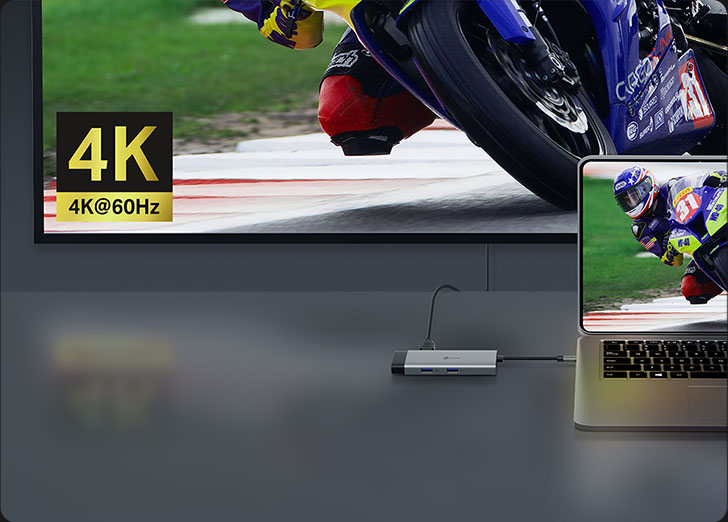Hub USB-C TP-Link UH6120C plateado conectado a una notebook que extiende la imagen de una carrera de motos en 4K@60Hz a un monitor o TV grande. El hub tiene puertos USB azules y un cable corto. Muestra la capacidad de visualización de alta resolución del producto.