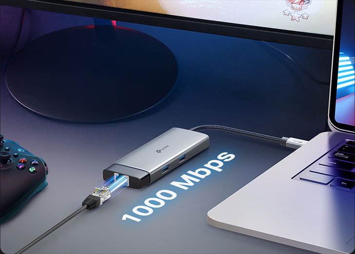 Hub USB-C TP-Link UH6120C conectado a una notebook con un cable Ethernet Gigabit (1000 Mbps) enchufado. El hub está sobre un escritorio oscuro junto a un control de videojuegos, con la notebook mostrando un juego de carreras. El texto 1000 Mbps sobre la imagen resalta la velocidad de conexión de red para juegos o trabajo.
