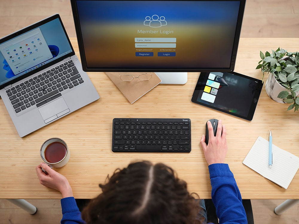 Vista aérea de una persona trabajando con el Teclado inalámbrico compacto Trust Lyra en un escritorio con dos computadoras, una tablet, una taza de café, un cuaderno con un bolígrafo y una planta. Una pantalla muestra la página de inicio de sesión del miembro con opciones para ingresar.
