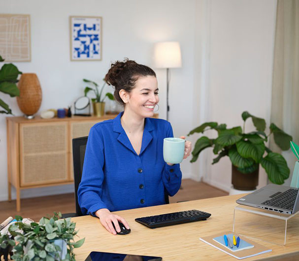 Una persona con una chaqueta azul sentada frente a un escritorio en una oficina en casa, sosteniendo una taza con la mano izquierda y operando un mouse con la derecha. Hay un portátil abierto, un Teclado inalámbrico compacto Trust Lyra color Negro. Al fondo se observa una estantería y una obra de arte abstracto en la pared.
