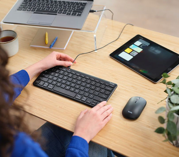 Una persona está sentada frente a una mesa de trabajo. En la mesa, hay un portátil abierto y una tableta digital encendida. La persona utiliza un Teclado inalámbrico compacto Trust Lyra color Negro y un mouse para interactuar con ambos dispositivos.