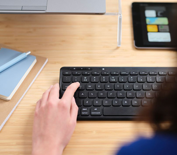 La imagen muestra una mano sobre un Teclado inalámbrico compacto Trust Lyra color Negro, presionando la tecla “Q”. Junto a la mano, hay una computadora portátil, una tableta y un cuaderno sobre un escritorio de madera.