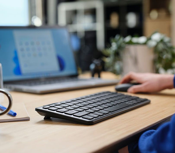 Primer plano de un escritorio con una mano utilizando un mouse, al lado de un Teclado inalámbrico compacto Trust Lyra color Negro y una laptop abierta.