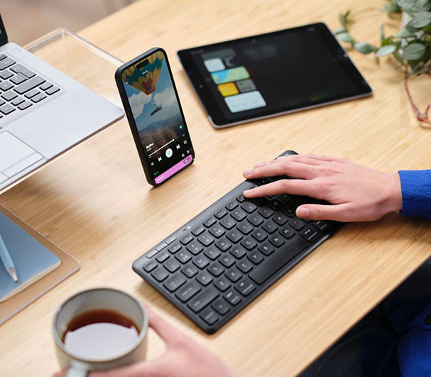 Imagen de una mesa de trabajo con un teléfono móvil apoyado en un soporte mostrando una imagen de globos aerostáticos en su pantalla, junto a una computadora portátil, una tableta digital y una persona utilizando el Teclado inalámbrico compacto Trust Lyra color Negro.