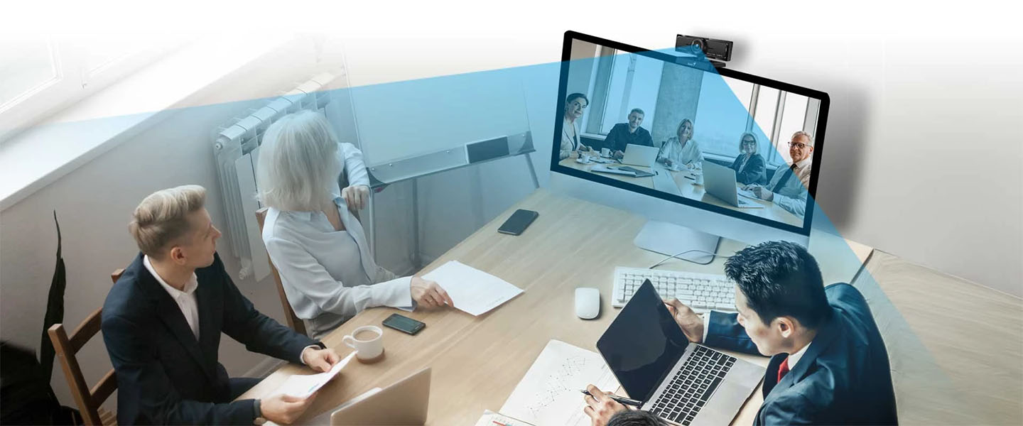 Webcam Genius WideCam F100 V2 capturando a tres personas en una videoconferencia en una sala de reuniones rectangular. El ángulo de visión ancho de la cámara en un monitor de pared captura a los participantes remotos, mientras que las tres personas en primer plano, con vestimenta de negocios, miran hacia la pantalla y trabajan en sus computadoras y documentos.