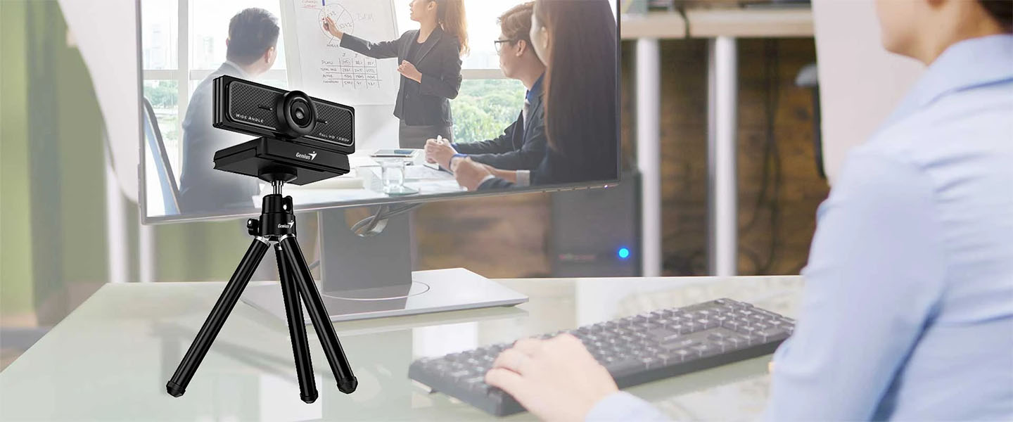 La webcam Genius WideCam F100 V2 está montada sobre un trípode pequeño sobre un escritorio, apuntando a una usuaria que trabaja en su computadora, vista desde atrás. En el monitor grande de la usuaria se ve una videoconferencia, con una mujer presentando gráficos a dos colegas en una oficina moderna.
