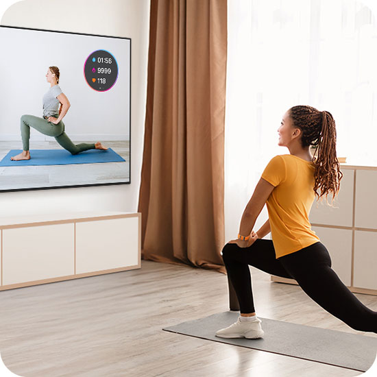 Una mujer realiza ejercicios de estiramiento frente a su televisor Crystal UHD, el cual muestra en pantalla a una instructora junto con un rastreador de entrenamiento en tiempo real que sincroniza sus datos de salud para que sigas tu progreso mientras entrenás.