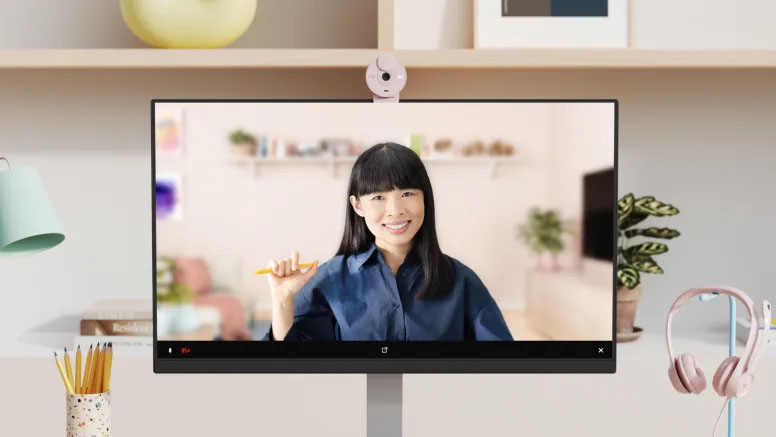 Una mujer sonriente en una videollamada. En el borde superior del monitor se ve una webcam Logitech Brio 300 de color rosa claro.