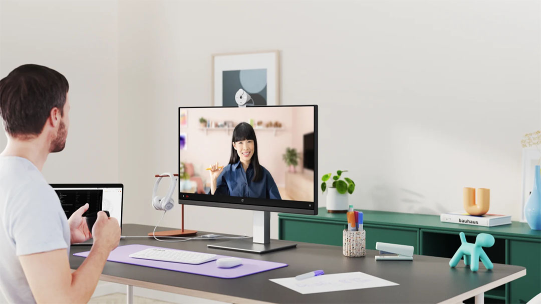 Un hombre en una videoconferencia en su escritorio con una webcam Logitech Brio 300 colocada encima de un monitor grande. Se ve a una mujer sonriente en la pantalla.