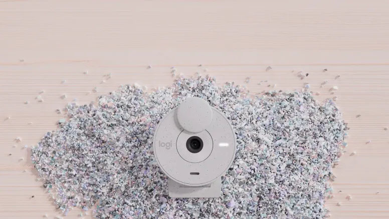 Una webcam Logitech Brio 300 de color blanco sobre una superficie de madera, rodeada de pequeños fragmentos de plástico reciclado.