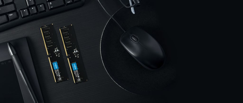 Dos módulos de memoria RAM Crucial DDR sobre un escritorio negro junto a un mouse, teclado y tableta gráfica