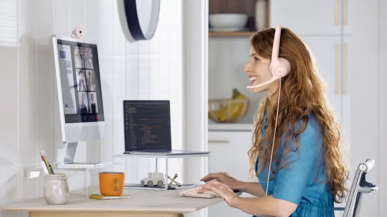 Mujer sonriendo usando auriculares Logitech H390 USB-C rosas en un escritorio de cocina para videollamadas, el mejor set para vos