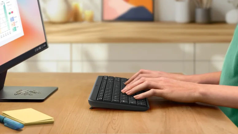 Persona usando el Teclado Compacto Inalámbrico Bluetooth Logitech K250 sobre un escritorio de madera junto a un monitor y notas adhesivas