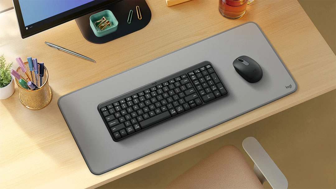 Teclado Compacto Inalámbrico Bluetooth® Logitech K250 con mouse sobre alfombrilla gris en escritorio de madera clara con monitor y accesorios de oficina