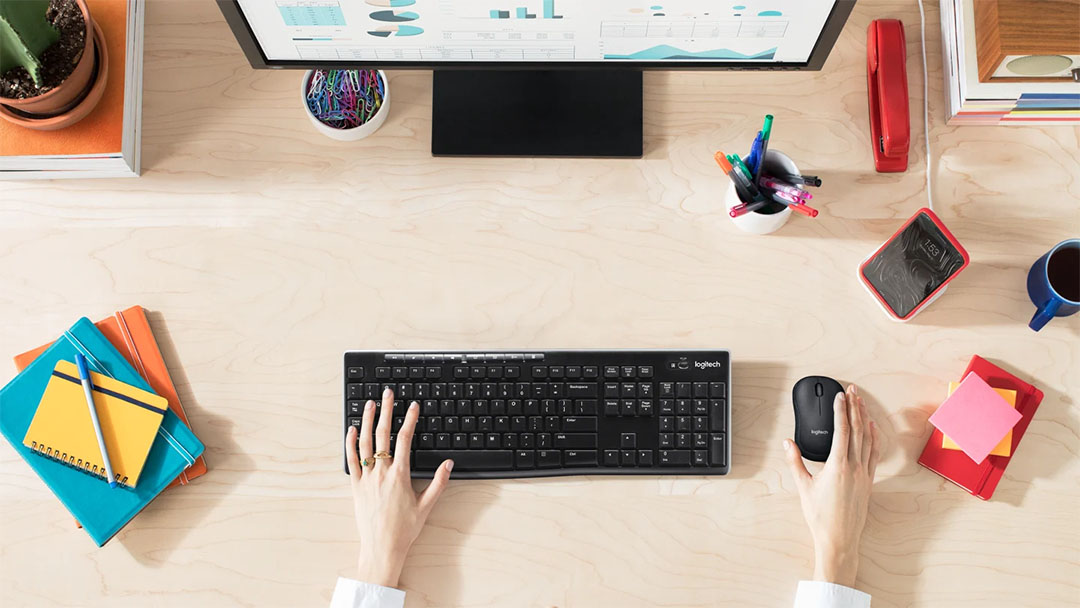 Persona usando el Teclado Inalámbrico Logitech K270 con mouse sobre un escritorio de oficina organizado, frente a un monitor con gráficos y rodeado de útiles de escritorio