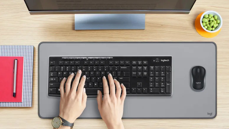 Persona usando el Teclado Inalámbrico Logitech K270 con mouse sobre alfombrilla gris en escritorio de madera, acompañado de monitor, cuaderno y recipiente con frutos secos