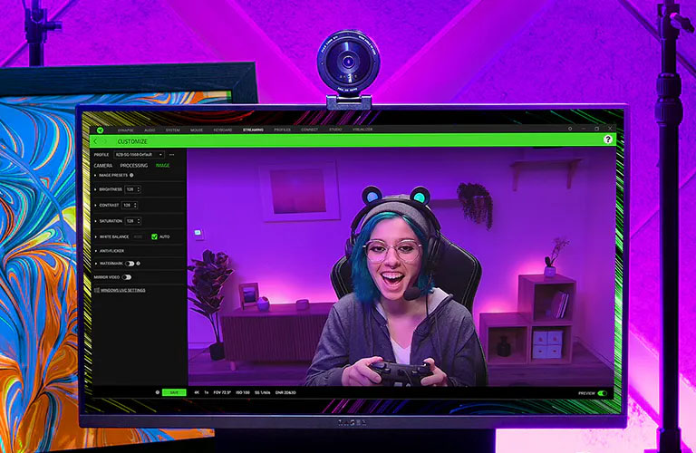 La imagen muestra una configuración de transmisión en vivo con iluminación de colores púrpura y azul. En el centro, hay un monitor que muestra a una persona con auriculares y un micrófono, sosteniendo un control de videojuegos. Encima del monitor, hay una cámara web Razer KIYO Pro. A la izquierda del monitor, hay un micrófono montado en un brazo ajustable, y a la derecha, unos auriculares con orejas de gato iluminadas. Detrás del monitor, se puede ver una pintura abstracta con colores vibrantes.