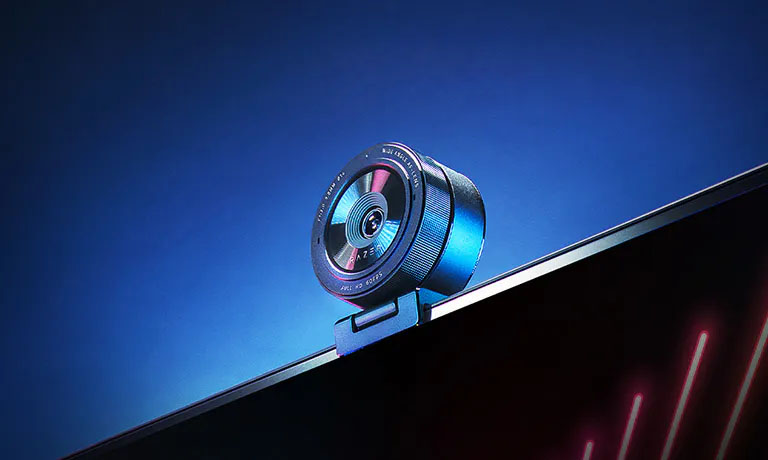 Una cámara web Razer KIYO Pro está montada en la parte superior de un monitor. La Razer KIYO Pro tiene un diseño circular, y se puede ver la lente en el centro. El fondo es de un color azul degradado, lo que resalta la cámara. El monitor muestra líneas diagonales de color rojo brillante.