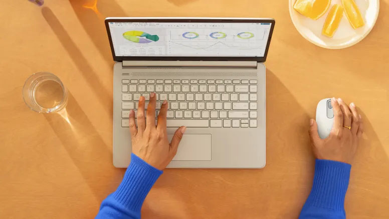 Persona usando el mouse Logitech M196 Bluetooth en color blanco junto a una notebook plateada sobre un escritorio con vaso de agua y plato con frutas