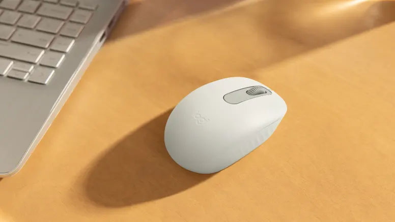 Mouse Logitech M196 Bluetooth en color blanco sobre un escritorio de madera junto a una notebook plateada