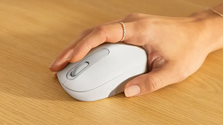 Mano usando el mouse Logitech M196 Bluetooth en color blanco sobre un escritorio de madera clara