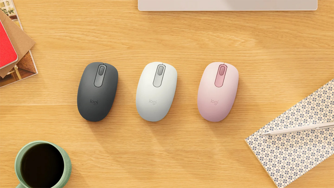 Mouse Logitech M196 Bluetooth en tres colores (grafito, blanco y rosa) sobre un escritorio de madera junto a taza, cuaderno y lapicera