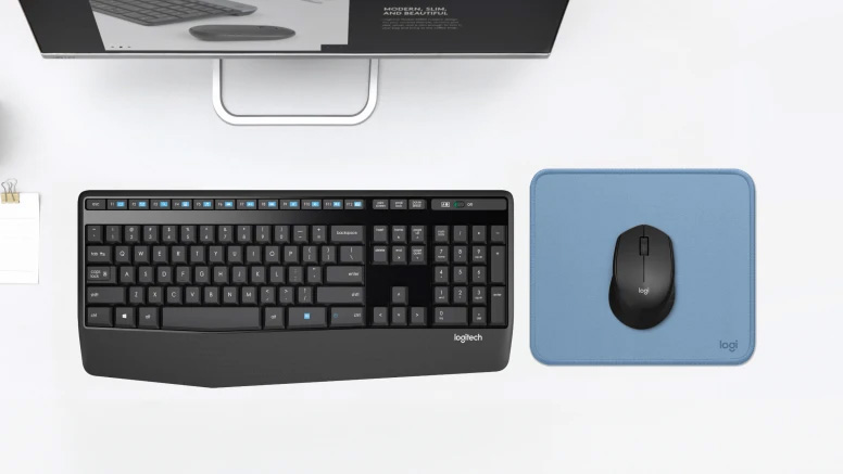 Vista superior de un escritorio con monitor, teclado Logitech negro y un Mouse Inalámbrico Logitech M280 de color negro sobre alfombrilla azul.