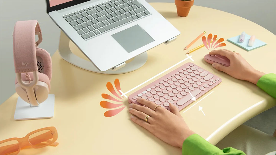 Teclado Logitech Pebble Keys 2 K380s rosado sobre un escritorio. Una persona está escribiendo y usando el mouse a juego. También se ve un portátil, auriculares y otros accesorios de oficina en tonos pasteles.