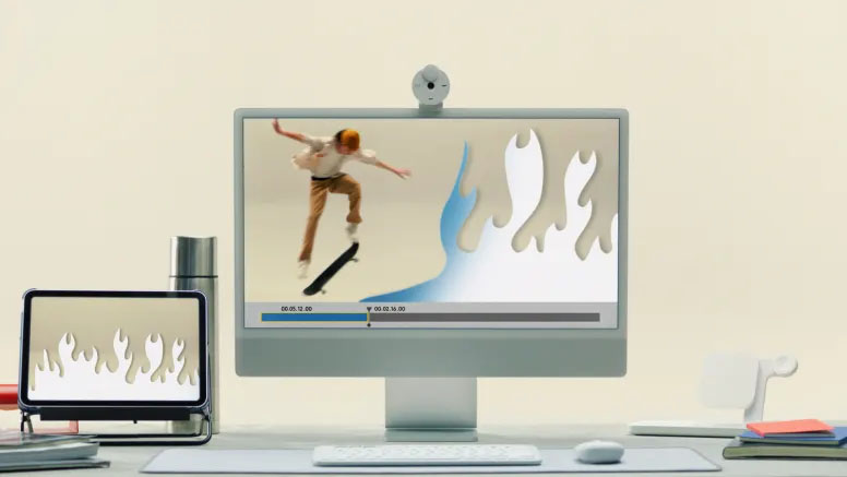 Una computadora de escritorio editando un video de un skater, al lado un iPad con un diseño de llamas, y en el escritorio se ve un teclado Pebble Keys 2 K380s blanco.