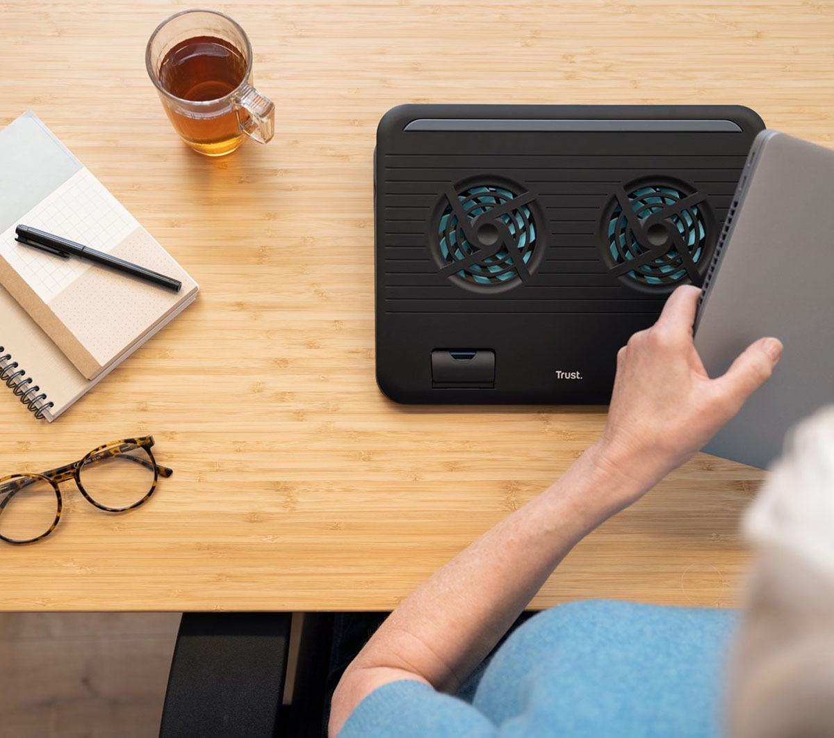 Persona utilizando una base de enfriamiento para portátil Trust Cyclone con dos ventiladores, sobre un escritorio de madera. Al lado, hay un cuaderno abierto, anteojos y una taza de té.