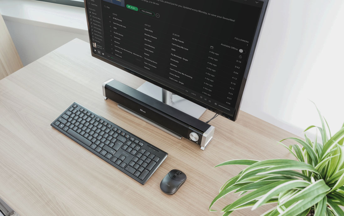 Monitor de computadora mostrando una lista de correos electrónicos con un teclado, un mouse y una Barra de sonido para PC y TV Trust Asto en un escritorio de oficina moderno.