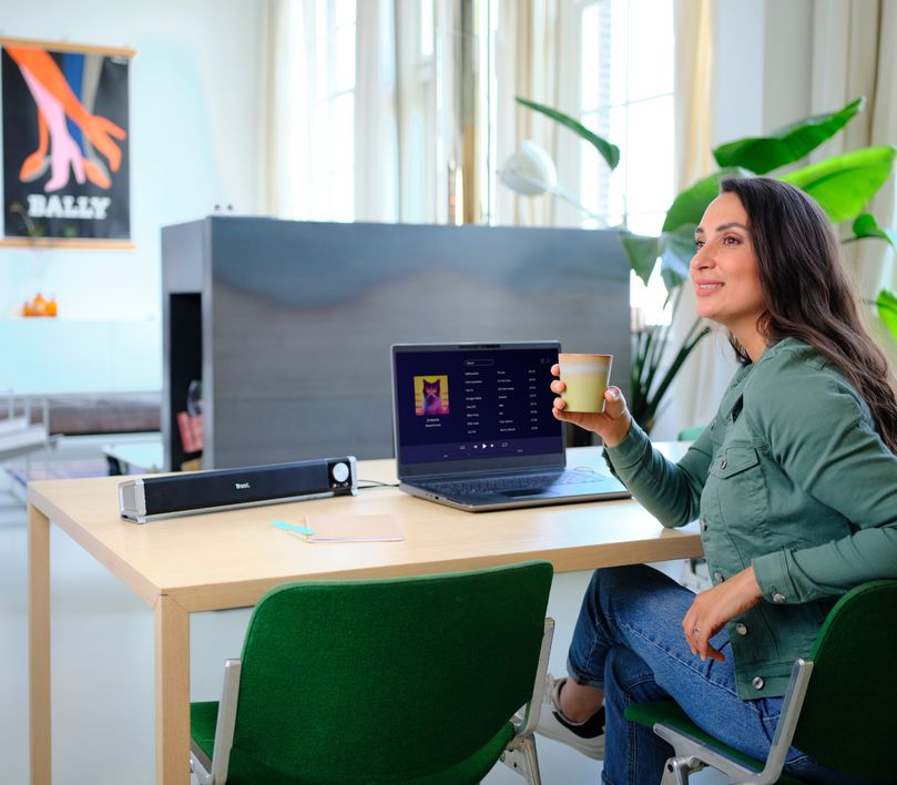 Una mujer está sentada frente a una laptop con gráficos en la pantalla, y una Barra de sonido para PC y TV Trust Asto conectada. Sostiene una taza.