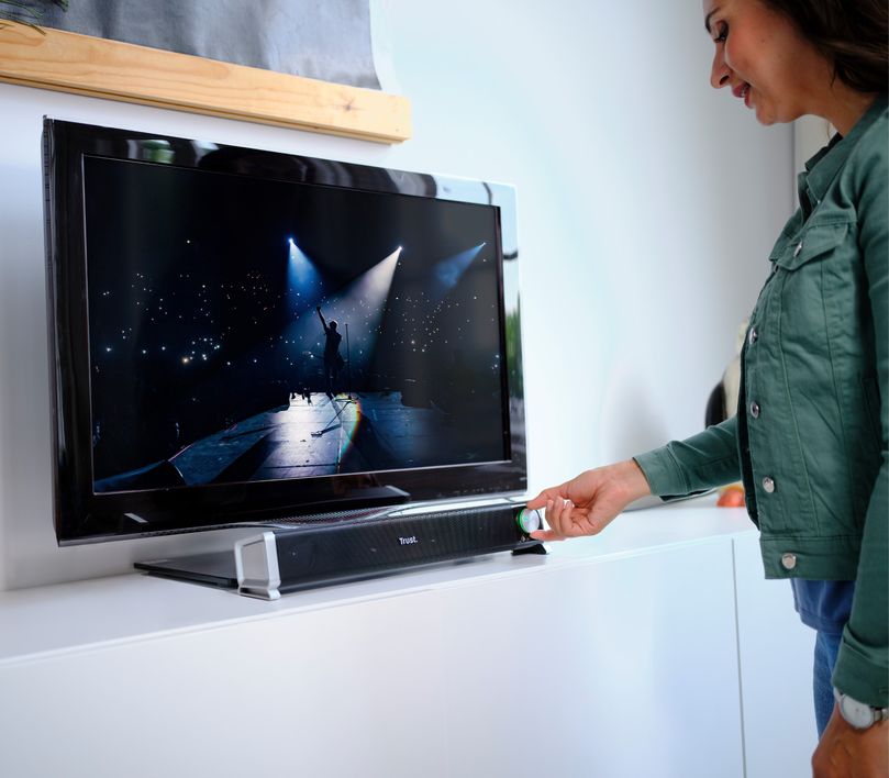 Una persona sostiene  el control de volumen de la Barra de sonido para PC y TV Trust Asto situada bajo un televisor de pantalla plana montado en la pared. En la pantalla del televisor se muestra una imagen de un conciero.