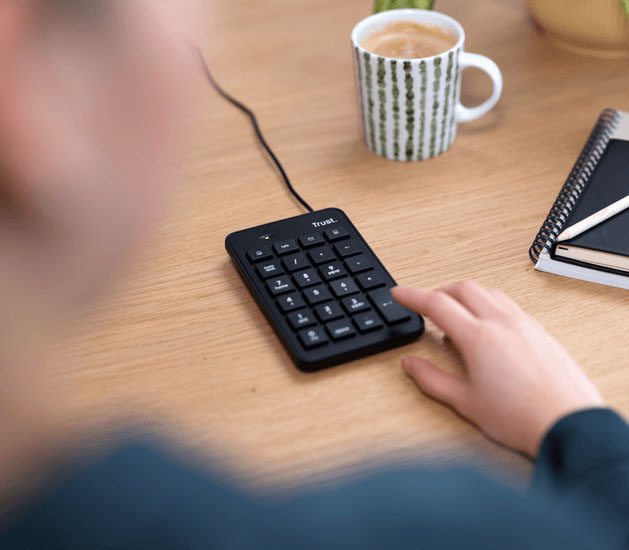Persona usando el teclado numérico USB Trust Xalas sobre un escritorio de madera, acompañado de una taza de café y una libreta con lápiz.