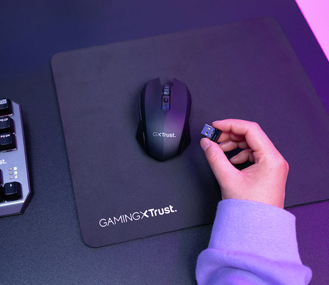 Mano sosteniendo el receptor USB inalámbrico de un Mouse gaming inalámbrico Trust GXT 115 Macci sobre un mousepad, con parte de un teclado.
