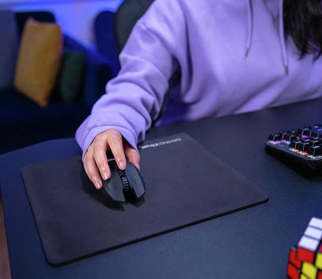 Mano usando un Mouse gaming inalámbrico Trust GXT 115 Macci inalámbrico sobre un mousepad oscuro, con parte de un teclado y un Cubo Rubik en el escritorio y luces de colores de fondo.