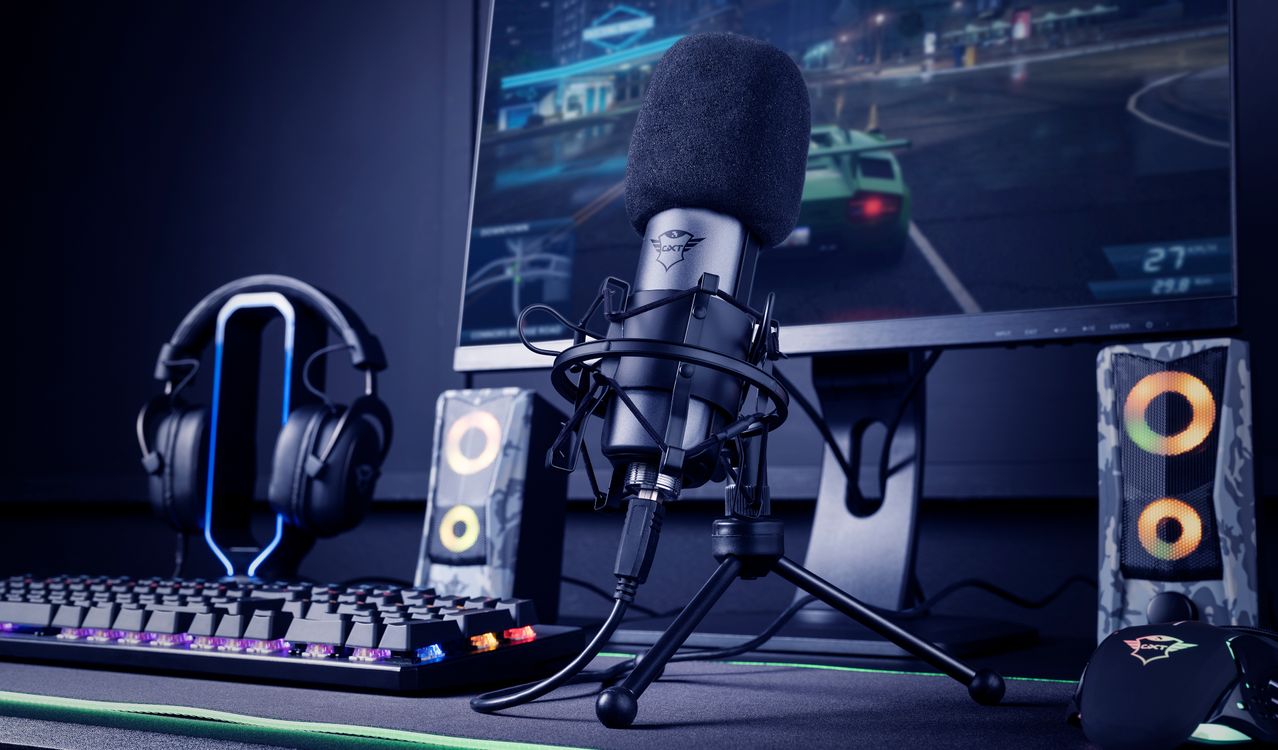 Un escritorio de gaming con un Micrófono USB para streaming Trust GXT 242 Lance en primer plano, teclado mecánico retroiluminado, auriculares de diadema, dos altavoces y un monitor mostrando un juego de carreras.