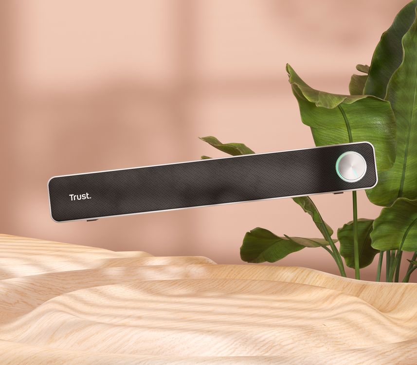 Barra de sonido para PC Trust Arys está flotando sobre una superficie de madera clara. Al fondo, se ven hojas verdes de una planta y un fondo difuminado en tonos rosados.