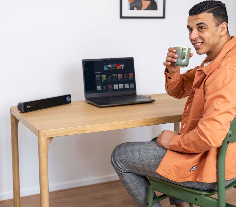 Una persona sentada frente a una computadora portátil sobre un escritorio de madera, sosteniendo una taza con la mano derecha. Hay una Barra de sonido para PC Trust Arys al lado izquierdo del escritorio.