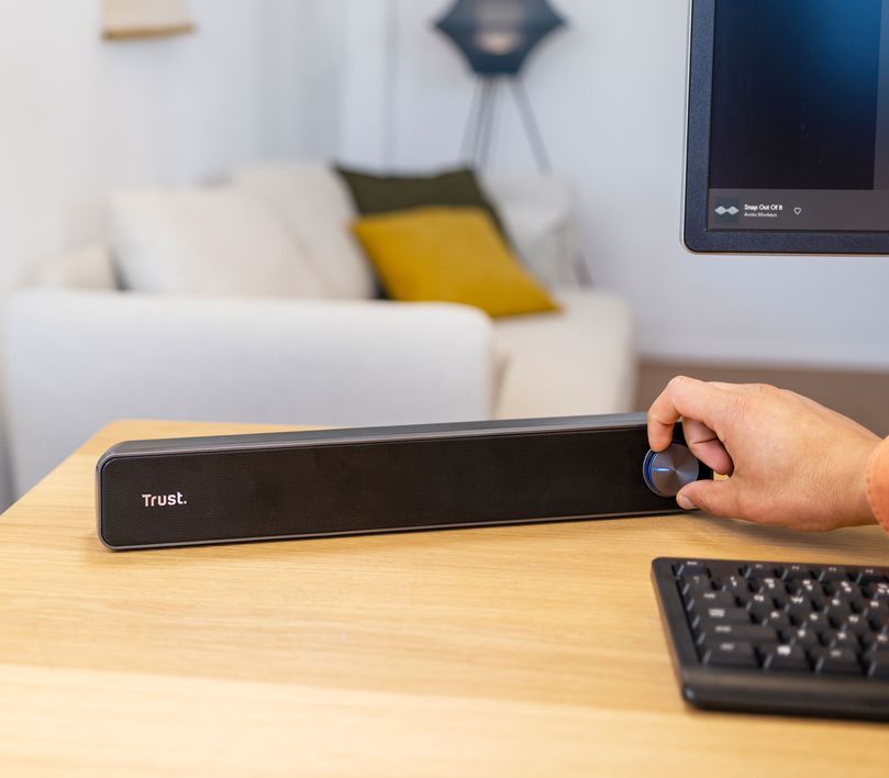 Una mano masculina ajustando el volumen de una Barra de sonido para PC Trust Arys en una oficina hogareña con un monitor, teclado y un sofá al fondo.