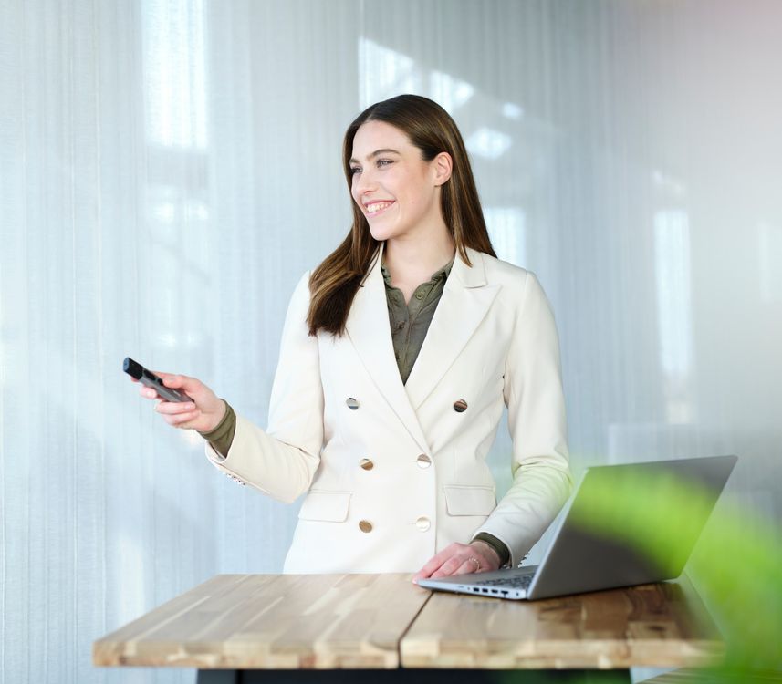 Mujer de negocios con traje blanco sentada frente a una laptop en un escritorio de madera, sosteniendo un teléfono Presentador inalámbrico con puntero láser Trust Kazun y mirando hacia la pantalla.