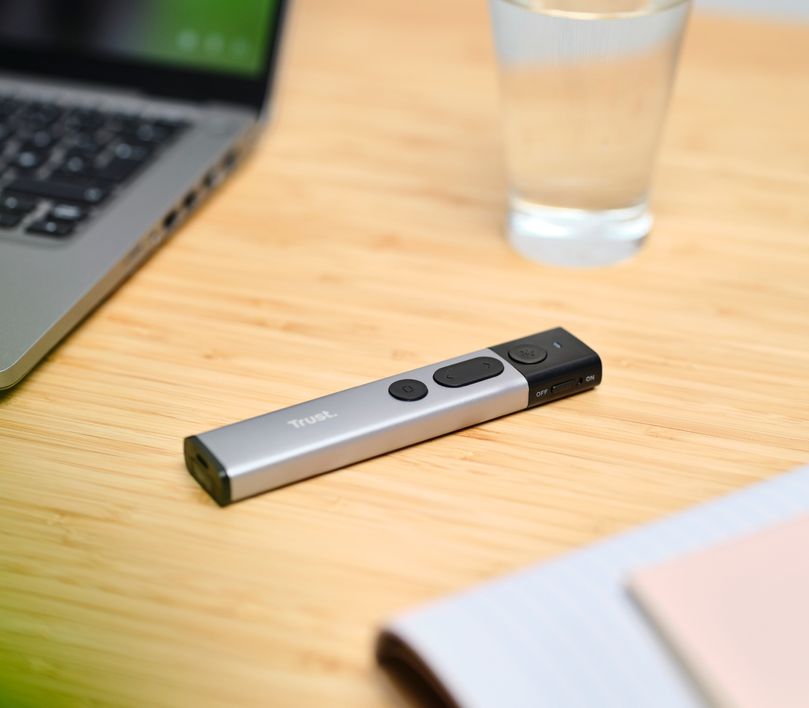 Presentador inalámbrico con puntero láser Trust Kazun sobre una mesa de madera junto a una computadora portátil, un vaso de agua y papeles parcialmente visibles.