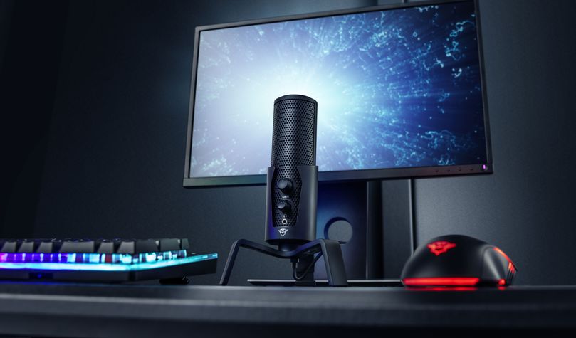 Imagen de un escritorio moderno con un monitor, teclado iluminado con luces RGB, un Micrófono USB 4 en 1 para streaming Trust GXT 258 Fyru y un mouse para juegos, todo sobre una superficie negra y contra un fondo oscuro.