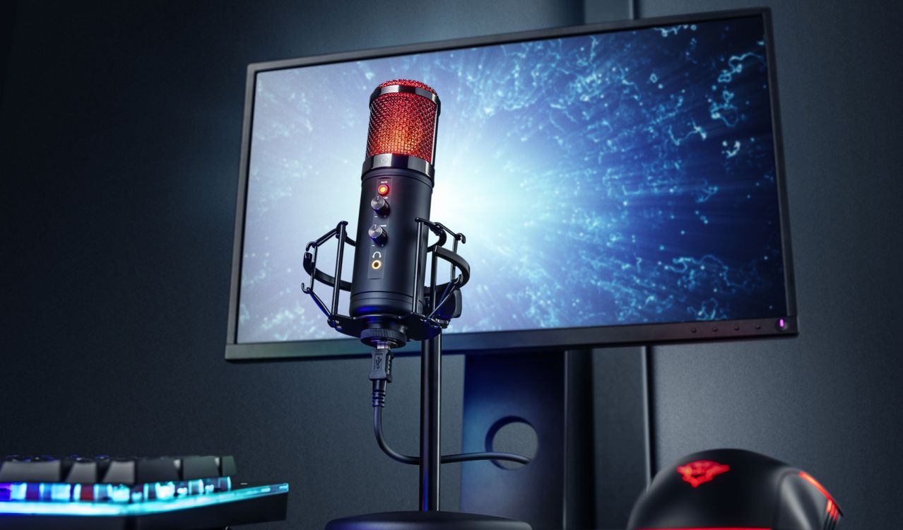 Micrófono Trust GXT 256 Exxo con luces rojas encendidas montado en un soporte frente a una pantalla de computadora que muestra una imagen abstracta de partículas azules, sobre un escritorio con teclado y mouse para juegos.
