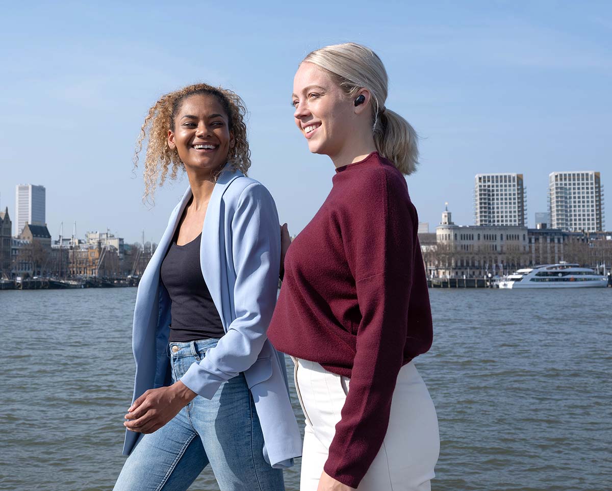 Dos personas caminando frente a un río con edificios en el fondo. Una de ellas tiene puestos unos Auriculares inalámbricos Bluetooth Trust Nika Compact color negro