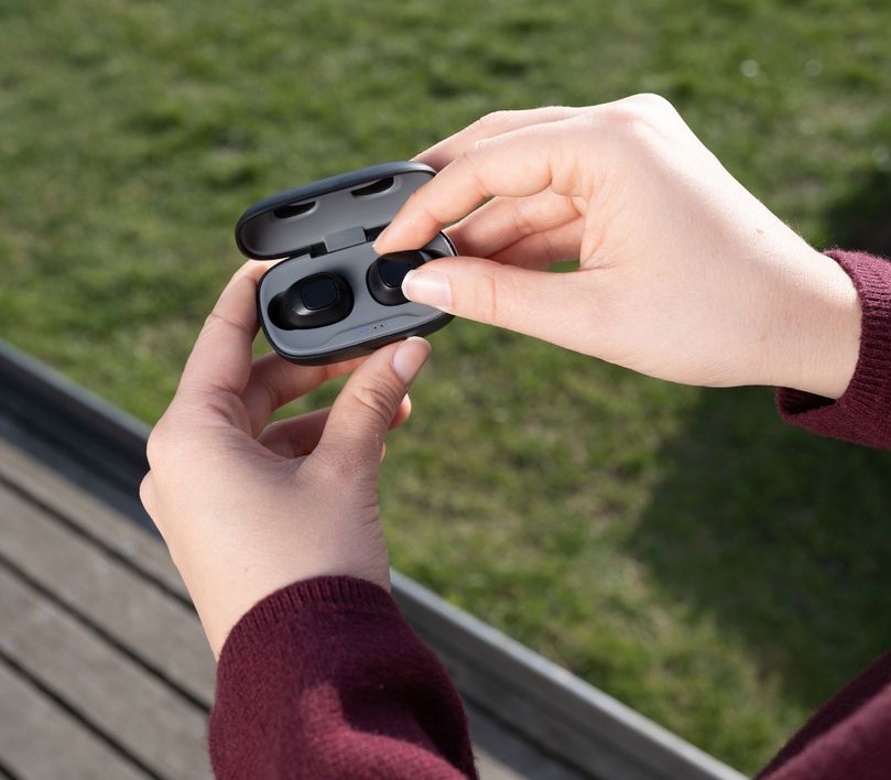 Una persona sosteniendo un estuche de carga abierto con Auriculares inalámbricos Bluetooth Trust Nika Compact color negro dentro, en primer plano, con un fondo desenfocado de césped verde.