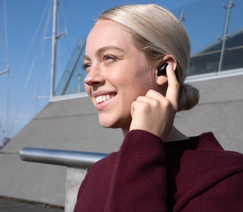 Persona con suéter color borgoña hablando por unos Auriculares inalámbricos Bluetooth Trust Nika Compact color negro, con el cabello rubio recogido y de fondo una barandilla metálica y mástiles de barcos desenfocados bajo un cielo despejado.