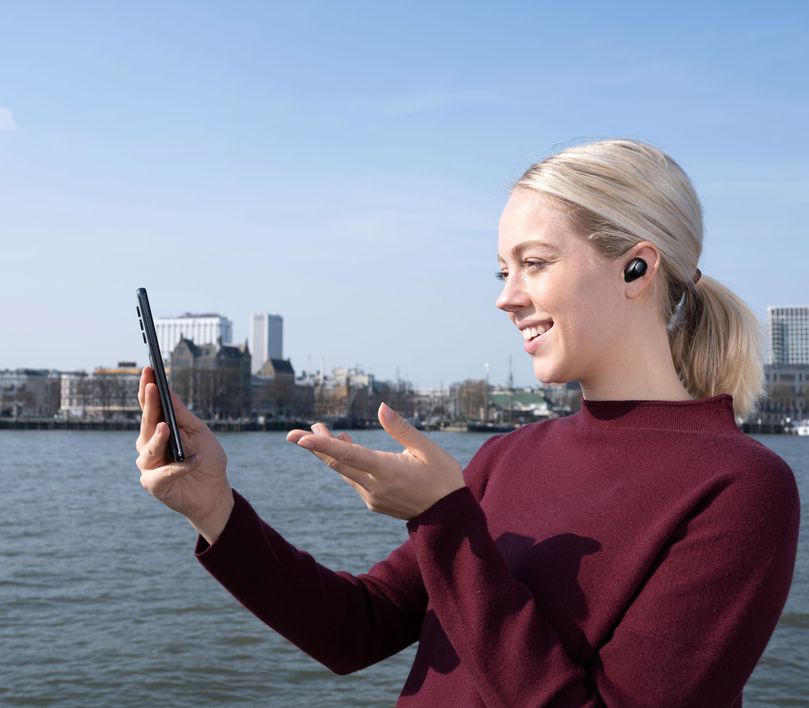 Una persona con cabello rubio, que lleva una remera roja y Auriculares inalámbricos Bluetooth Trust Nika Compact color negro, sostiene un teléfono inteligente frente a un cuerpo de agua con edificios al fondo.