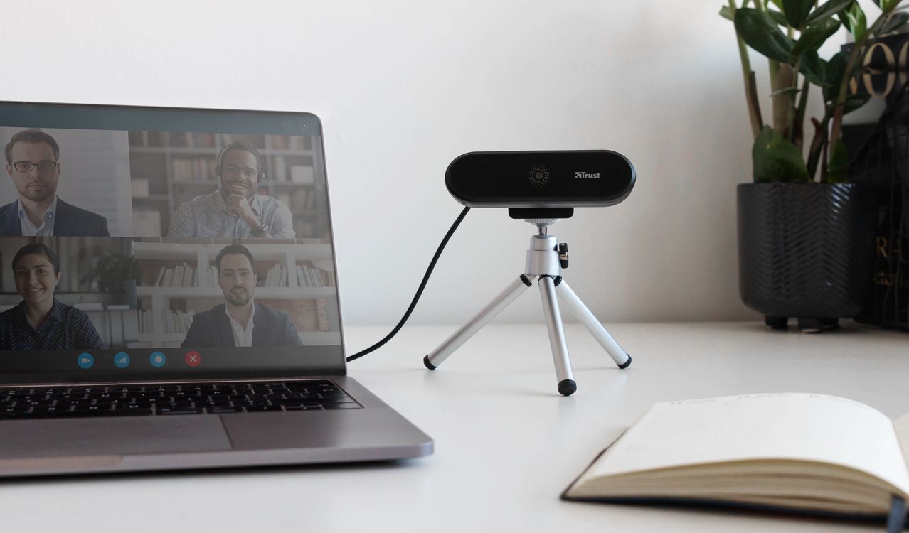 En la pantalla de una computadora portátil, se muestra una videollamada grupal con tres participantes. Una Cámara web Full HD Trust Tyro está colocada en un trípode cerca de la computadora, y hay un cuaderno abierto en primer plano a la derecha. La escena se desarrolla sobre un escritorio con fondo blanco y algunas decoraciones, como una planta.